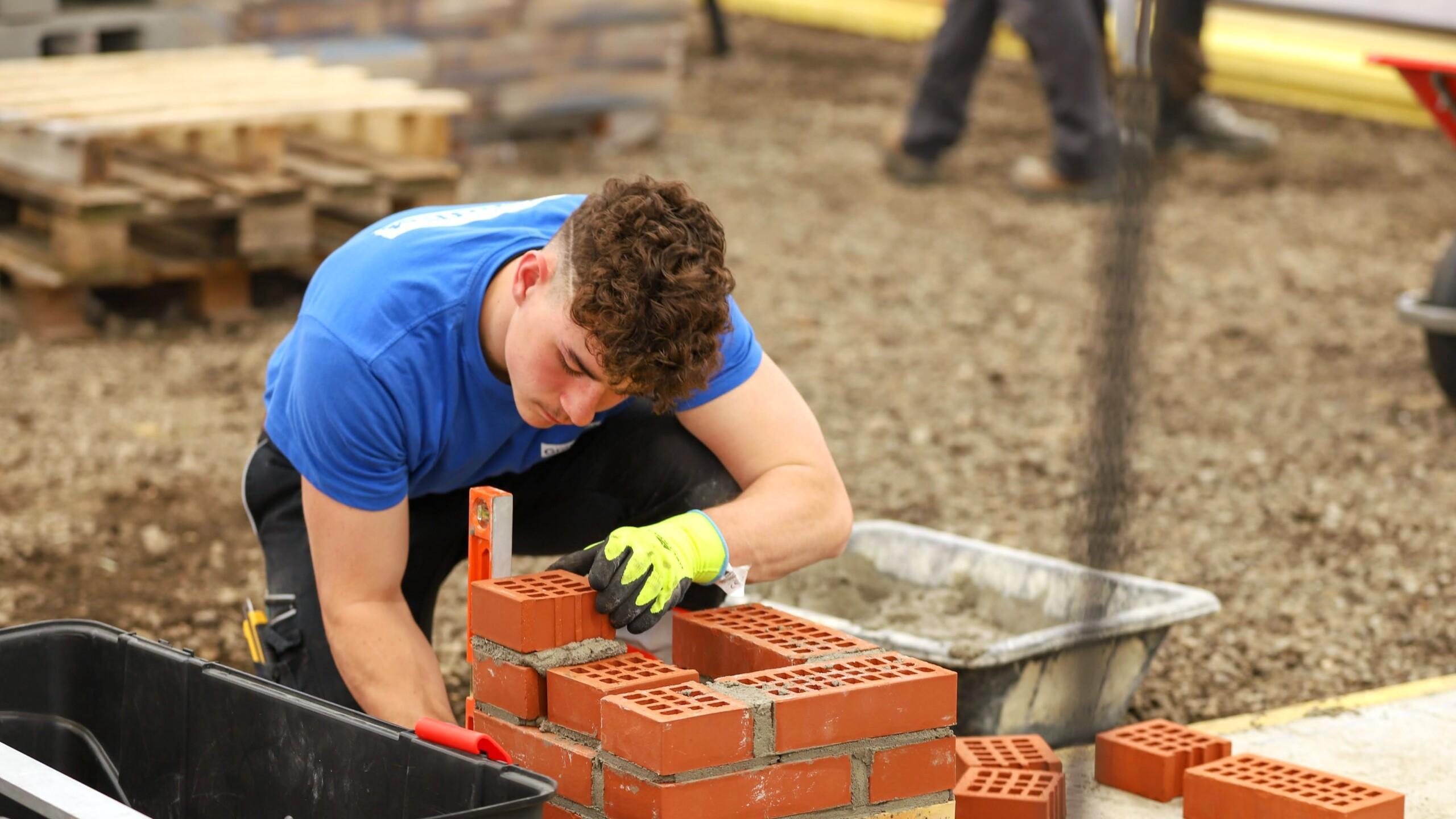 A young competitor in a blue shirt and yellow gloves carefully lays bricks during a bricklaying skills competition.